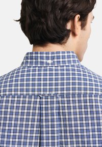 Camisa de manga corta, con botones y un patrón de cuadros en azul y blanco. Presenta un cuello estándar y un plisado en la parte posterior para ajustar el ajuste.
