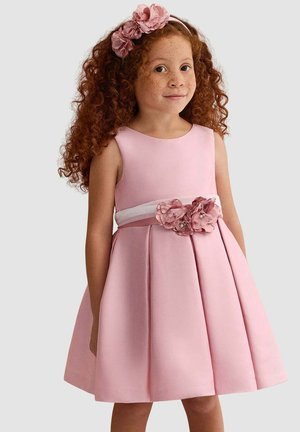 Ragazza giovane con capelli ricci rossi che indossa un vestito rosa senza maniche con fascia floreale per capelli e cintura decorata con fiori abbinati.