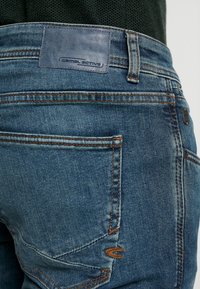 Dunkelblaue Denim-Jeans mit einer verblassten Textur. Verfügt über ein Lederpatch mit der Aufschrift "camel active" und kontrastierendem orangefarbenem Nähten an den Taschen.