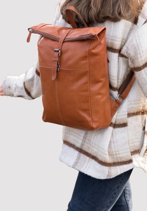 Person, der eine weiß-braun karierte Jacke trägt und einen großen braunen Leder-Rucksack mit Schnalle und Reißverschluss auf dem Rücken trägt.