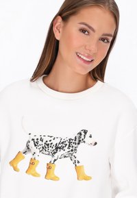 Fehér pulóver egy sárga gumicsizmát viselő dalmata kutya grafikájával. A mintán fekete foltok vannak a kutyán, és egyszerű, kerek nyakkivágással rendelkezik.