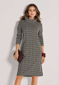 Eine Frau trägt ein knielanges Kleid mit langen Ärmeln in schwarz-beiger geometrischer Musterung, hält eine dunkle geflochtene Clutch und trägt rote Armreifen.
