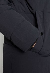 Chaqueta acolchada en tejido azul marino oscuro con una superficie texturizada. Presenta un bolsillo oculto y un cierre con botones negros para un aspecto elegante.