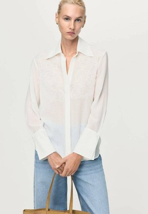 Blusa bianca con motivo floreale ricamato, maniche lunghe e chiusura a bottoni sul davanti. Tessuto morbido e leggero con una leggera trasparenza.