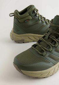 Zielone buty trekkingowe sięgające do połowy kostki, z gumową podeszwą, teksturowanym materiałem i syntetycznymi wstawkami. Oferują płaskie sznurowadła i wzmocnioną piętę dla lepszego wsparcia.