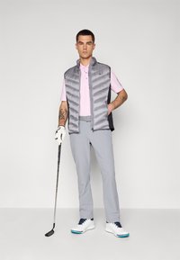 Sivi puffer prsluk s visokim ovratnikom, ispod kojeg je svijetloružičasta polo majica, svijetlosive hlače, bijele golf cipele i golf palica u ruci.