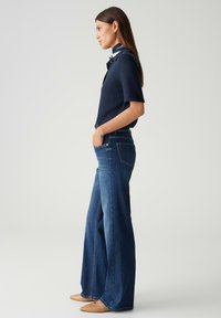 Marineblaues Kurzarmblusenshirt mit Knopfdetails, kombiniert mit weiten, dunklen Jeans. Das Model trägt nacktfarbene, spitz zulaufende Ballerinas mit schlichten Design.