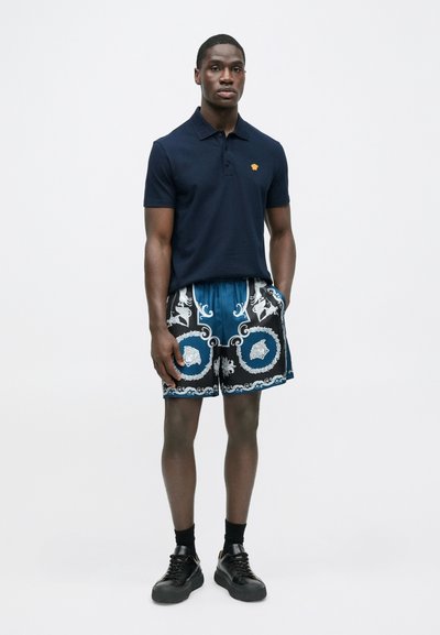 Μπλε πόλο πουκάμισο με κίτρινο λογότυπο, σε συνδυασμό με μπλε μαγιό με σχέδια. Το μοντέλο φοράει μαύρα αθλητικά παπούτσια και κάλτσες μέχρι τους αστραγάλους.