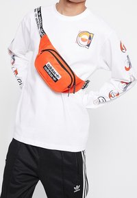 Orange Adidas waist bag with two zippered compartments, accented with a black strap featuring white Adidas branding. White long-sleeve shirt worn.