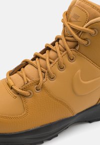 Zapatilla alta de cuero marrón y malla con ojales reforzados, suela negra contrastante y acentos de tela texturizada. Logo de Nike en el lateral exterior.