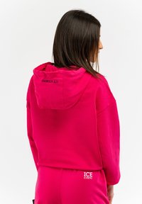 Felpa fucsia con cappuccio, cordino e dettaglio del logo sul cappuccio. Tessuto morbido, vestibilità oversize, abbinata a pantaloni coordinati.