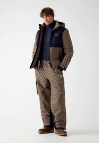 Καφέ σετ μπουφάν και παντελόνι με σχέδιο και ναυτικές λεπτομέρειες. Το υλικό έχει υφή με ανάγλυφη επιφάνεια· διαθέτει φερμουάρ, κουκούλα και τσέπες.