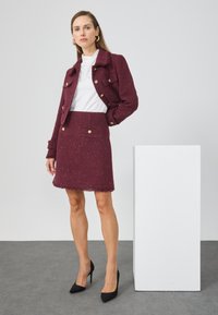 Burgundijas krāsas tweed jaka un svārku komplekts ar zeltītām pogām, saplēsušiem malām un teksturētu apdari, valkāts ar melniem augstpapēžu apaviem.