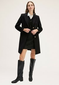 Černý dlouhý kabát se zlatými knoflíky, nošený přes texturované černé šaty. Modelka v černých vysokých botách, stojící na neutrálním pozadí.