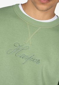 Felpa verde con ricamo di "Harper" in un font corsivo. Il materiale sembra morbido con colletto a costine e dettagli di cucitura.