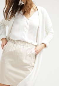 Cardigan en tricot blanc sur une blouse blanche légèrement translucide, associé à une jupe beige à taille élastique avec poches, texture lisse.