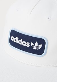 Hvit caps med et marineblått rektangel som har ordet "adidas" i hvitt og et trefoil-logo, kantet med lys blå søm.