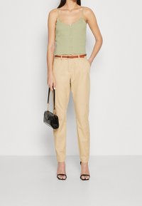 Groen geribd tanktop met knoopdetail, beige taps toelopende broek, bruine riem, zwarte handtas en zwarte sandaalhakken op een lichte achtergrond.