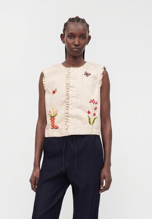 Kvinde med fletninger iført ærmeløs beige top med blomster- og sommerfuglebroderi, kombineret med løse mørke bukser mod en ensfarvet baggrund.