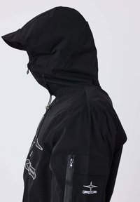 Persona che indossa una giacca nera con cappuccio, dotata di una tasca con zip sul braccio e un'etichetta bianca con il logo "PXP" sulla manica, rivolta a sinistra.