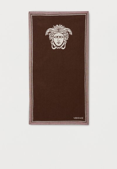 Καφέ ορθογώνια πετσέτα με λευκό έμβλημα κεφαλής Μέδουσας κοντά στο πάνω κέντρο και τη λέξη "VERSACE" γραμμένη στην κάτω δεξιά γωνία.
