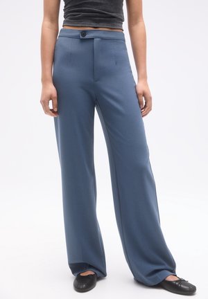 Femme portant un pantalon bleu taille haute à jambes larges, associé à des ballerines noires et un haut noir côtelé court.