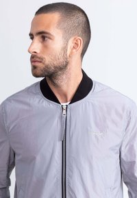 Giacca bomber grigia chiara con colletto a coste nero, chiusura con zip e dettaglio ricamato sul petto. Texture liscia, design minimalista.