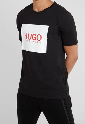 Man wearing black T-shirt with large white square and red "HUGO HUGO BOSS" logo on the front, paired with black pants.
