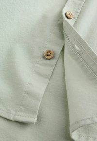 Camicia di cotone verde chiaro con una texture liscia, caratterizzata da una patta con bottoni e bottoni in legno. Cuciture rifinite e dettaglio del colletto piegato visibili.