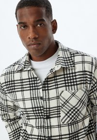 Camisa de franela a cuadros en blanco y negro con cuello abotonado, bolsillo frontal y camiseta gris debajo. Textura suave y patrón a cuadros.