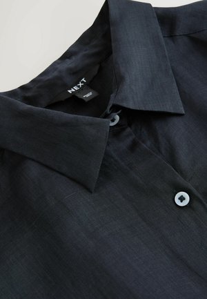 Tumši zila pogām aiztaisāma krekls ar gludu tekstūru, tai ir apkakle un neliels balts pogas dekors uz pogas ailes.