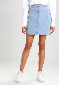 Valkoinen denimhame vaaleansinisessä sävyssä, tiukalla vyötäröllä, suoran leikkauksen ja valkoisilla ompeluominaisuuksilla. Yhdistettynä valkoisiin lenkkareihin.