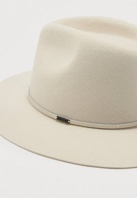 Chapéu fedora de feltro bege claro com uma fita fina a condizer e um pequeno logótipo da Brixton metal no lado da coroa.