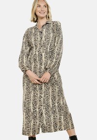 Usmívající se žena s blonďatými vlasy, oblečená v šatech s dlouhým rukávem, knoflíky a potiskem hadí kůže, a černých kotníkových botách na bílém pozadí.