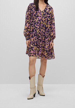 Kobieta ubrana w luźną sukienkę z długim rękawem i dekoltem w kształcie litery V, ozdobioną abstrakcyjnym wzorem w kolorach fioletowym, czarnym i złotym, zestawioną z beżowymi botkami na obcasie do połowy łydki.