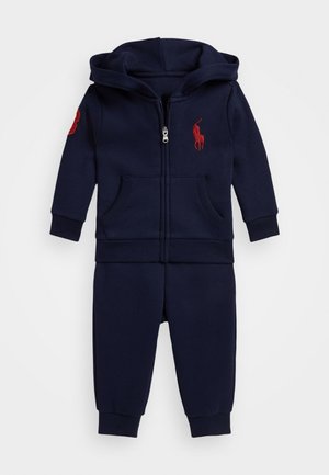 Námornícky modrý teplákový komplet z bavlny, obsahujúci mikinu na zips s prednými vreckami, rebrované manžety a červené vyšívané logo na hrudi.