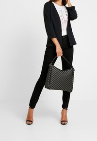 Borsa intrecciata nera con una forma strutturata, dotata di manico corto e chiusura con zip. Abbinata a un blazer nero, jeans neri e tacchi.