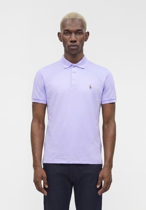 Homme aux cheveux blonds courts portant un polo violet clair à manches courtes et un pantalon foncé, debout devant un fond blanc.
