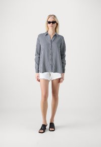 Camisa a rayas blanca y negra con cierre de botones, combinada con pantalones cortos blancos deshilachados y mules negros de punta abierta, con un fondo neutro.