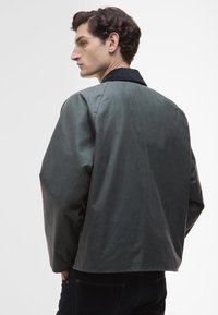 Γκρι, ακατέργαστο καμβά σακάκι με μαύρο γιακά, χαλαρή εφαρμογή, κατεβασμένοι ώμοι και λεία υφή. Ορατή ραφή στο πίσω μέρος.