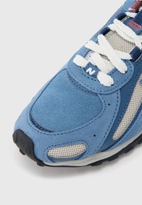 Μπλε αθλητικό παπούτσι σουέντ με πλέγμα, λευκά κορδόνια και μαύρη λασ�τιχένια σόλα. Διακριτικό λογότυπο 'N' στη γλώσσα, με λεπτομέρειες ραφής.