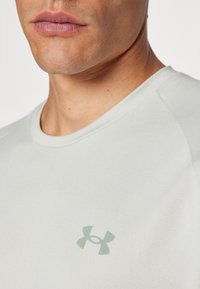 Maglietta a manica corta grigio chiaro con colletto rotondo, realizzata in tessuto liscio, con un sottile logo verde di Under Armour sul petto.