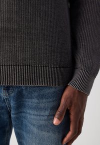 Pull en tricot gris foncé avec une texture côtelée et des poignets, associé à un jean en denim bleu. Une main visible repose sur le jean.