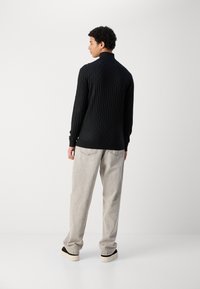 Schwarzer gerippter Pullover mit hohem Kragen und langen Ärmeln, kombiniert mit hellgrauen, locker sitzenden Hosen und schwarzen Schnürschuhen.