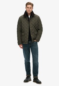 Giacca trapuntata verde oliva con colletto in velluto a coste marrone, chiusura a scatto e due tasche anteriori, abbinata a jeans blu e stivali neri.