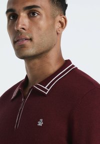 Burgunderfarbener Halbreißverschluss-Pullover mit kontrastierendem, weiß gestreiftem Kragen, gestricktem Muster und aufgesticktem Pinguin-Logo auf der Brust.