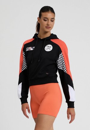 Felpa cropped nera e arancione con accenti a quadri, dotata di logo frontale, cappuccio con cordini e polsini a coste. Indossata con pantaloni corti arancioni.