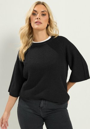 Lyshåret kvinde iført en sort ribstrikket sweater med 3/4-ærmer og mørke jeans, poserer mod en ensfarvet lys baggrund.