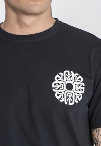 Camiseta de algodón negra con cuello redondo que presenta un diseño gráfico blanco en la parte izquierda del pecho, mostrando símbolos entrelazados.