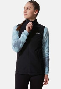 Schwarze ärmellose Jacke mit zwei Seiten Taschen, getragen über einem hellblauen Batik Langarmshirt; verfügt über einen hohen Kragen und einen Frontreißverschluss.